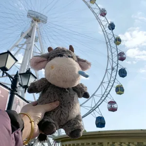 Cute grey cow plush doll held by hand in front of a giant Ferris wheel under a clear blue sky