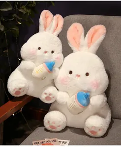 Close-up of two soft white plush bunnies holding blue baby bottles in a warm indoor setting