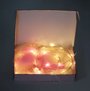 Caja de regalo atrapasueños con plumas rosas y luces cálidas en papel triturado blanco.