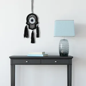 Hand-woven Hamsa Evil Eye wall hanging with black tassels in a minimalist interior setting.