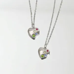 Elegante collar de corazón plateado con pedrería de colores sobre fondo de espejo.