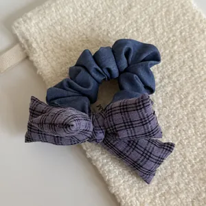 Close-up detail of a purple plaid bow scrunchie displayed on a cream textured pouch