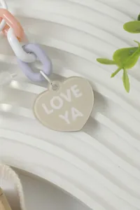 Close-up detail of a beige heart-shaped charm with LOVE YA text on a rainbow chain.