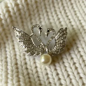 Elegante broche de doble cisne con pedrería y perla sobre fondo de punto.