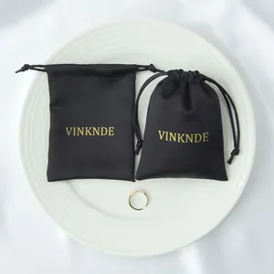 Bolsas de regalo de satén negro con cordón y logotipo dorado sobre un plato de cerámica blanca.