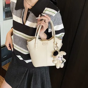 Trendy women's cream bucket handbag with cute plush bear charm and lychee texture.