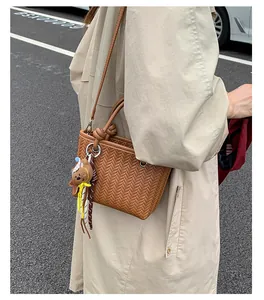 Detailansicht der braunen gewebten Beuteltasche mit Bärenanhänger am beigen Trenchcoat.