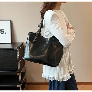 Large black shiny tote bag carried by a model in a minimalist interior setting.