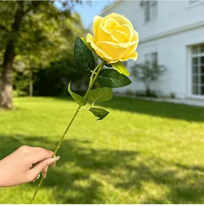Close-up of a single vibrant yellow artificial rose held against a sunny garden backdrop.