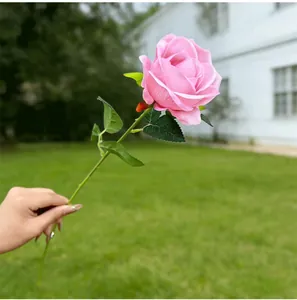 Handheld single pink artificial rose flower with realistic green stem and leaves outdoors