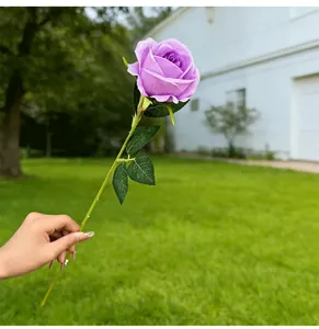 Hand holding a detailed purple artificial rose stem against a lush garden backdrop.