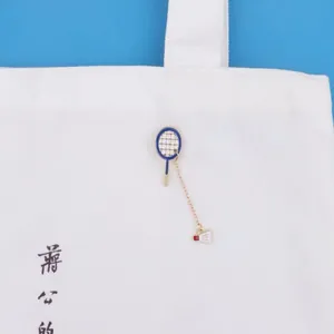 Detailed view of a badminton racket enamel pin with a gold chain on a white canvas tote bag.