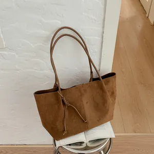 Minimalist large brown suede tote bag for women leaning against a white textured wall on a stool