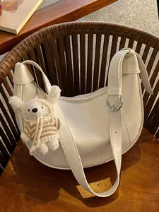 Stylish white crescent crossbody bag with cute plush bear charm on wooden chair. Perfect gift.