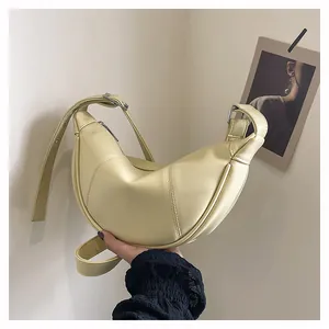 Hand haelt eine blassgelbe Croissant-Schultertasche vor neutraler Wand