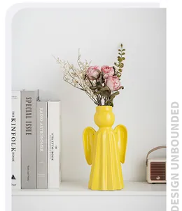 Modern yellow angel shaped ceramic vase with pink dried roses on a white shelf.