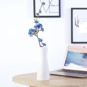 Elegant minimalist white ceramic flower vase with blue artificial flowers on a wooden desk.