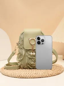 Bolso bandolera verde con colgante de gato y perla, mostrado junto a un móvil para escala.