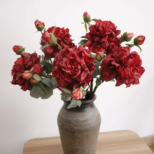 Realistic artificial red silk peony flowers in a textured ceramic vase on a wooden table.