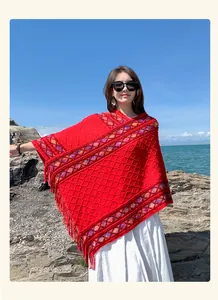 Ethnic red knitted poncho with decorative tassels worn by model at the rocky seaside
