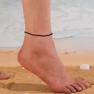 Bracelet de cheville minimaliste à perles noires sur un pied nu sur une plage de sable.