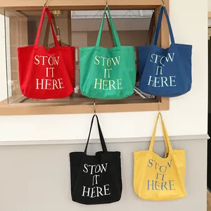 Borse tote in tela colorate con stampa 'STOW IT HERE' appese a ganci su una parete chiara.