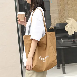 Large brown nylon tote bag with white letter print carried by a model in a casual setting.