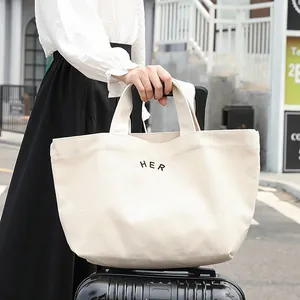 Bolso tote de lona blanca grande con texto 'HER' sostenido por una mujer en viaje