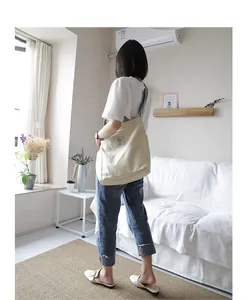 Lifestyle view of woman carrying a large beige canvas crossbody bag in a cozy room