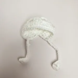 Close-up of white handmade knitted mini doll hat with ties and decorative pom-poms