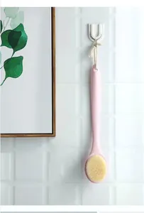 Pink long handle bath brush with soft bristles hanging on a white tiled bathroom wall.