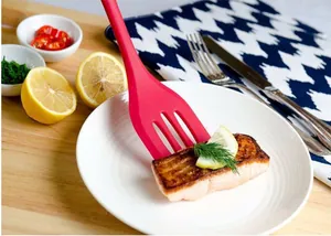 Close-up view of a red silicone kitchen spatula lifting a grilled fish fillet on a white plate