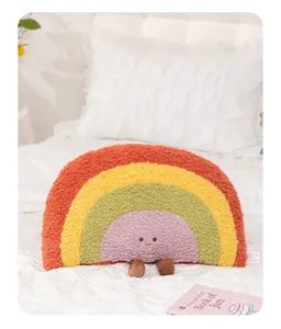 Soft rainbow plush pillow with cute face and dangling legs placed on a white cozy bed