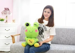 Close-up of cute green cartoon frog plush toy with heart held by person on sofa