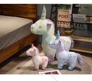 Three pastel rainbow unicorn plush toys in various sizes arranged in a cozy bedroom setting.