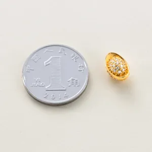 Makroaufnahme einer kleinen goldenen Yuanbao-Perle mit Pavé-Kristallen neben einer Münze.