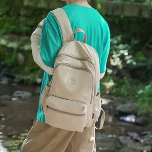 Detail shot of a casual beige nylon backpack worn by a person in a scenic forest stream setting.