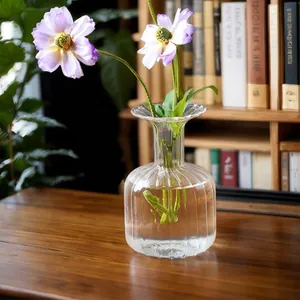 Clear ribbed glass vase with purple flowers on a wooden desk with a bookshelf background.