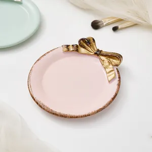 Detalle de un plato de cerámica rosa para joyas con un lazo dorado y borde texturizado.