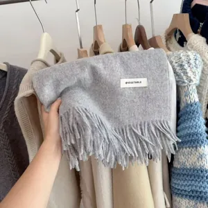Close-up detail of a thick grey knit scarf featuring fringes and a minimalist brand label.