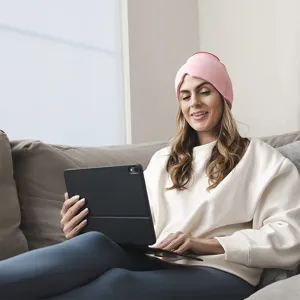 A woman relaxing on a sofa wearing a pink gel cooling headgear while using her tablet