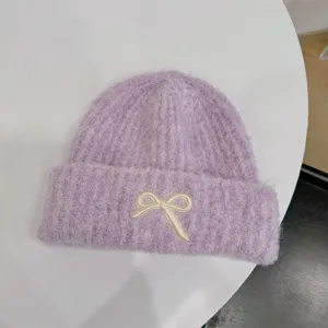 Detail of a purple fuzzy beanie with an ivory embroidered bow on the folded cuff.