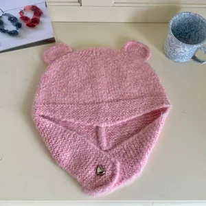 Detailed view of pink knitted bear-ear beanie with silver heart button on cream desk