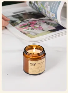 Close-up view of a lit scented soy wax candle in an amber glass jar on white marble surface