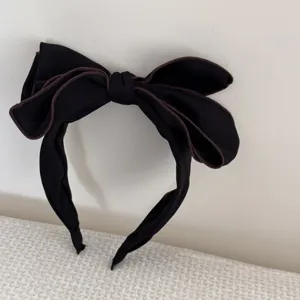 Primo piano di un cerchietto nero con fiocco 3D oversize e dettagli di bordatura marroni.