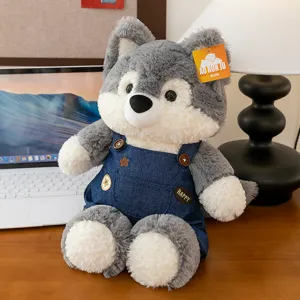 Cute husky plush toy wearing denim overalls sitting on a wooden desk with a laptop