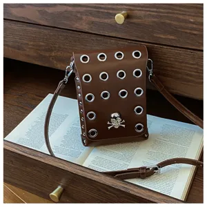 Brown skull crossbody bag with silver eyelets and rivets on a vintage book backdrop.