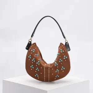 Brown crescent hobo bag decorated with turquoise stones and gold studs on white podium