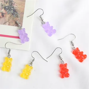 Pendientes de ositos de acrílico translúcido en morado, amarillo y rojo sobre tela blanca.