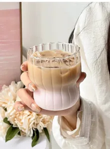 Hand holding a ribbed glass cup with layered coffee and milk in a soft light setting.